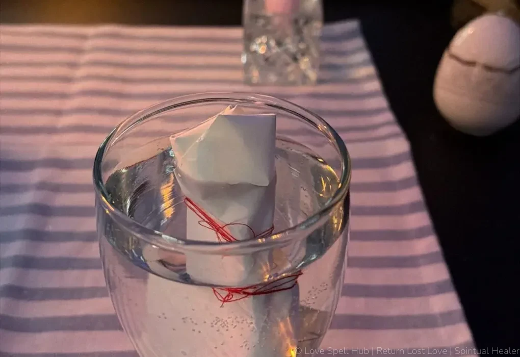 A folded petition paper immersed in a glass of water next to a burning pink candle and a white spiritual pot for cleansing rituals